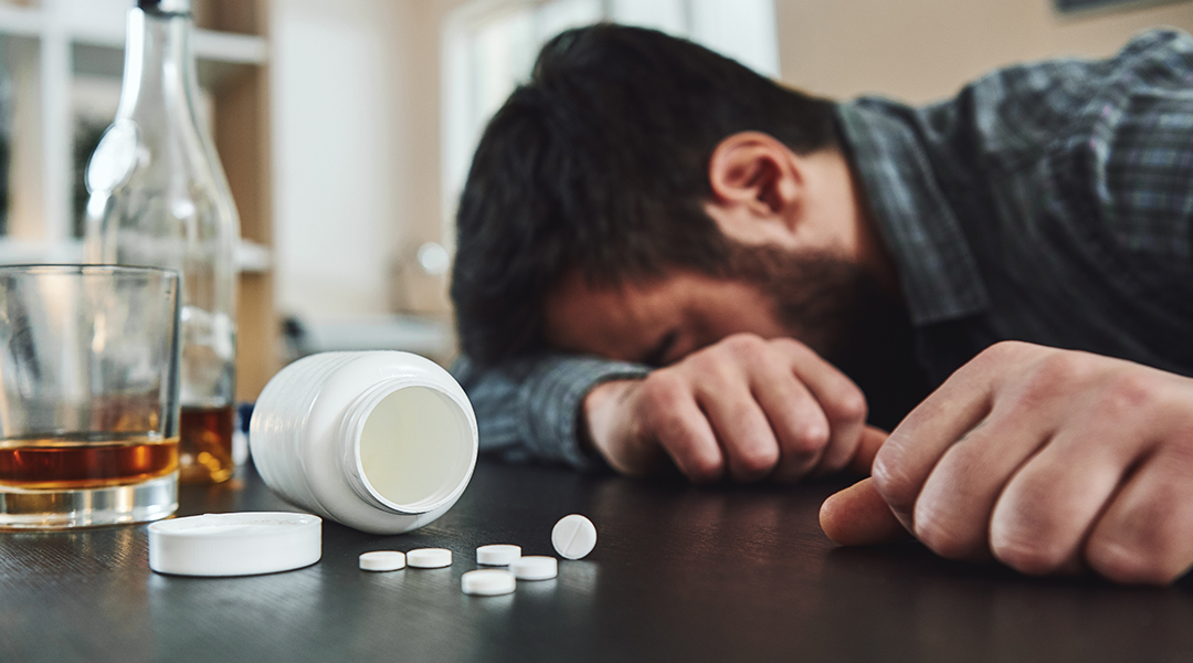 man laying with his head on arm with open bottle of pills and alcohol