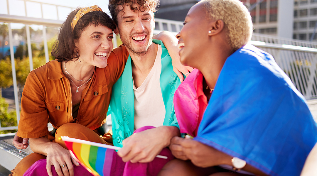 Three friends smiling and chatting outdoors on a sunny day, with a rainbow Pride flag visible, celebrating together.