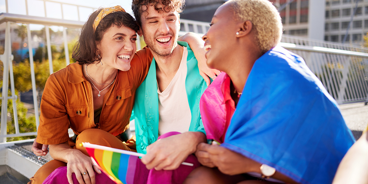 Three friends smiling and chatting outdoors on a sunny day, with a rainbow Pride flag visible, celebrating together.