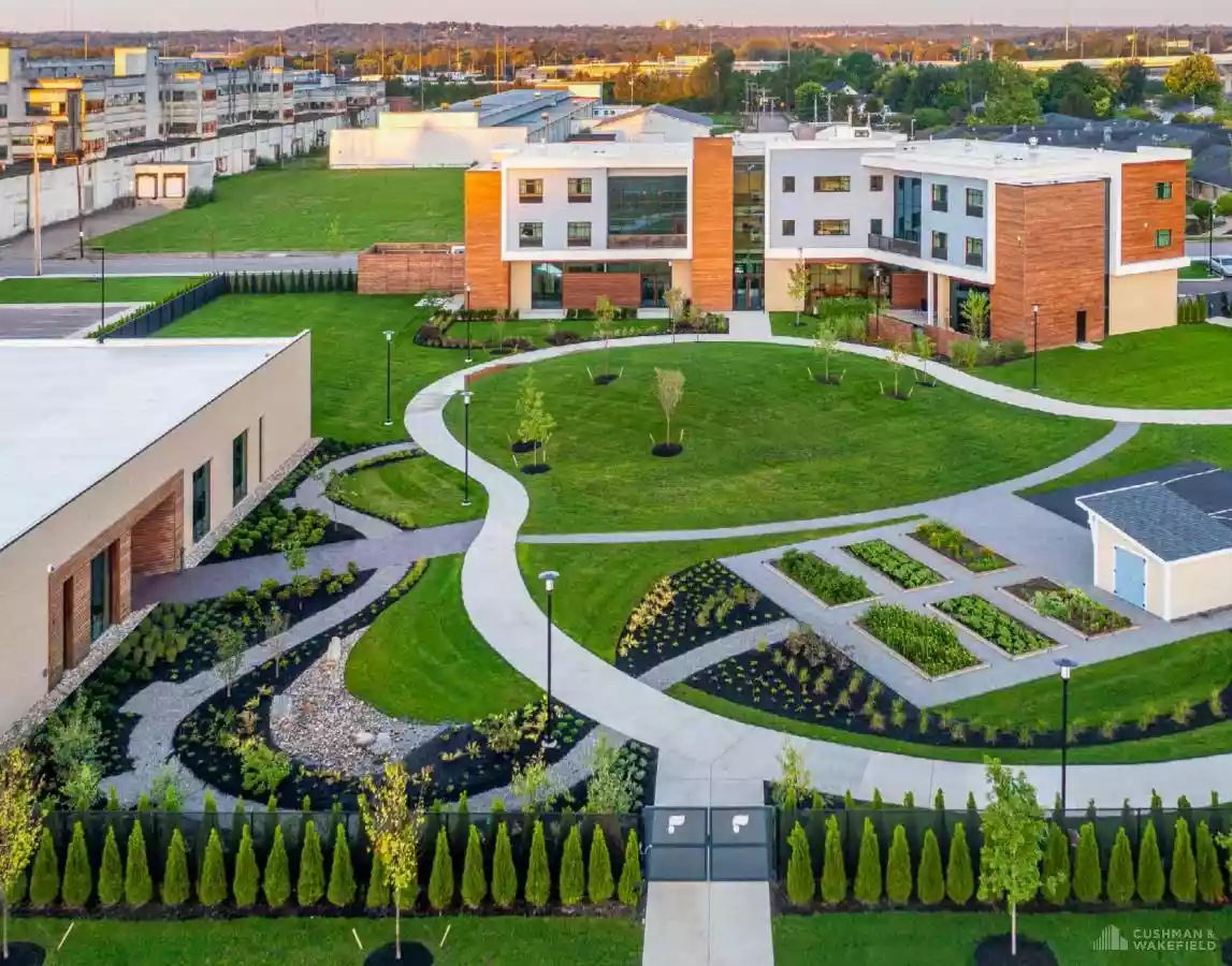 Aerial view of a modern campus with curved concrete paths, lush green lawns, and white-and-brick buildings arranged around a central courtyard garden area.