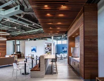 Open-plan coworking lounge with a kitchen island, bar stools, and scattered tables and chairs under a wood ceiling.