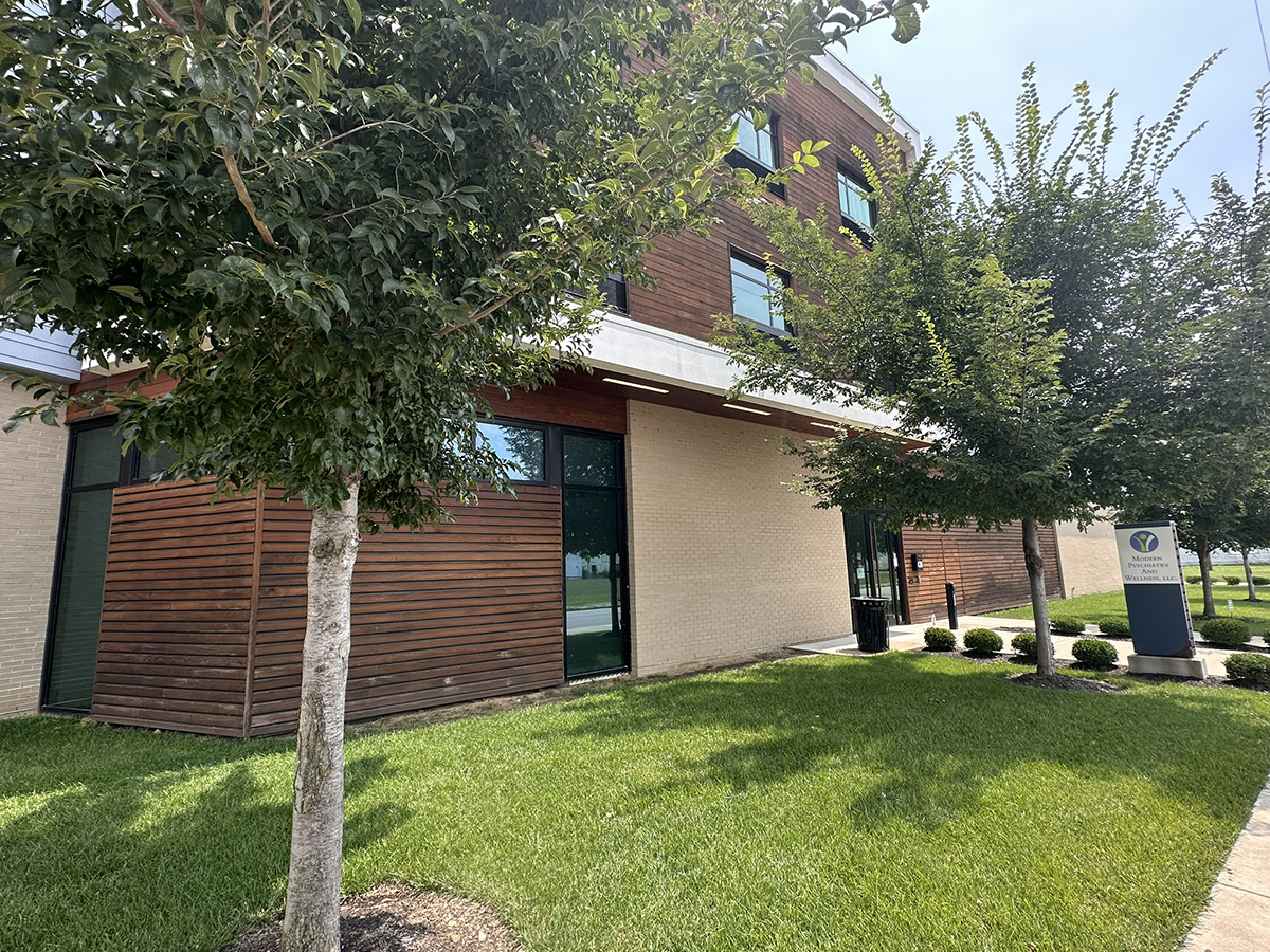 Modern office building with wood-panel siding, large windows, and a well-kept lawn with trees along the walkway.