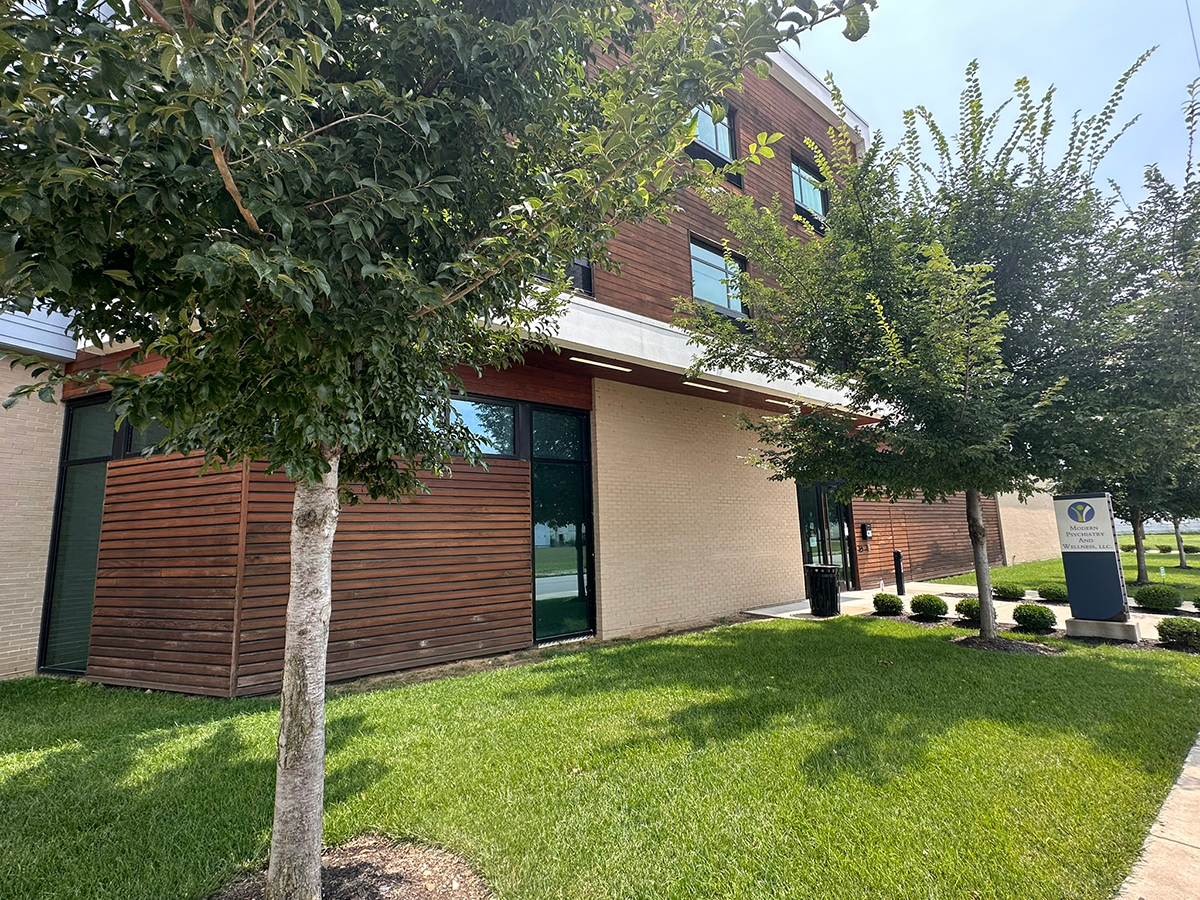 Front of a modern brick and wood-clad office building with a green lawn and two trees along the walkway; entrance area visible with a sign nearby.