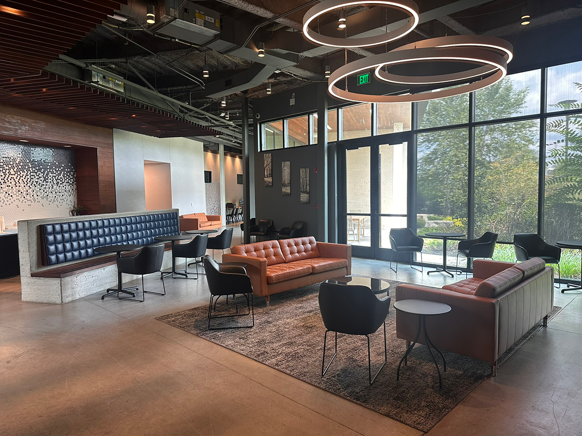 Modern lounge area in a contemporary lobby with blue tufted seating, orange sofas, black chairs, circular pendant lights, and large windows looking out to greenery.