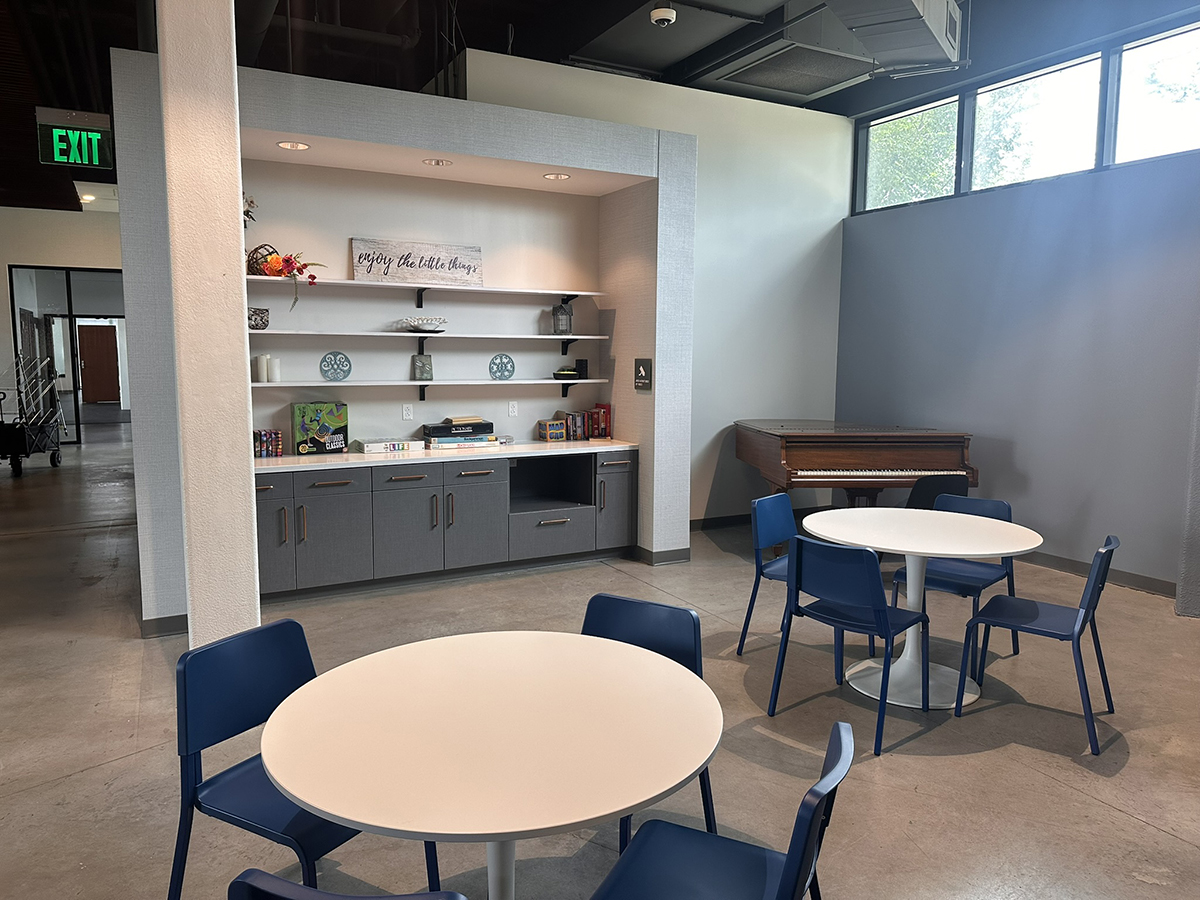 Modern open lounge area with round white tables and blue chairs, a built-in shelving unit with decorative items, and a piano against a gray wall; green Exit sign visible at left.