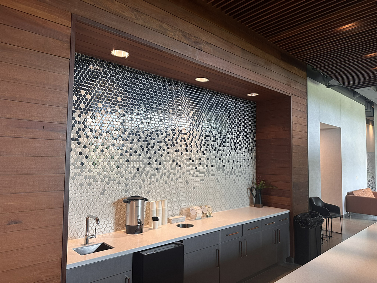 Modern beverage station with wooden wall panels, hex tile backsplash, and gray cabinetry under a white countertop; coffee machine and cups on the counter.