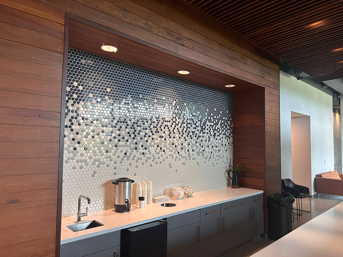 Self-serve coffee station in a lobby: white countertop, dark gray cabinets, hex tile backsplash, coffee urn, cups, sink, and a plant.