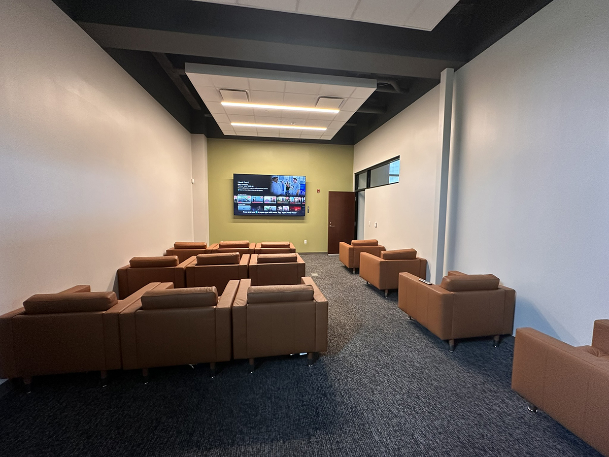 Meeting lounge with brown leather couches arranged in rows facing a wall-mounted TV on a green accent wall.