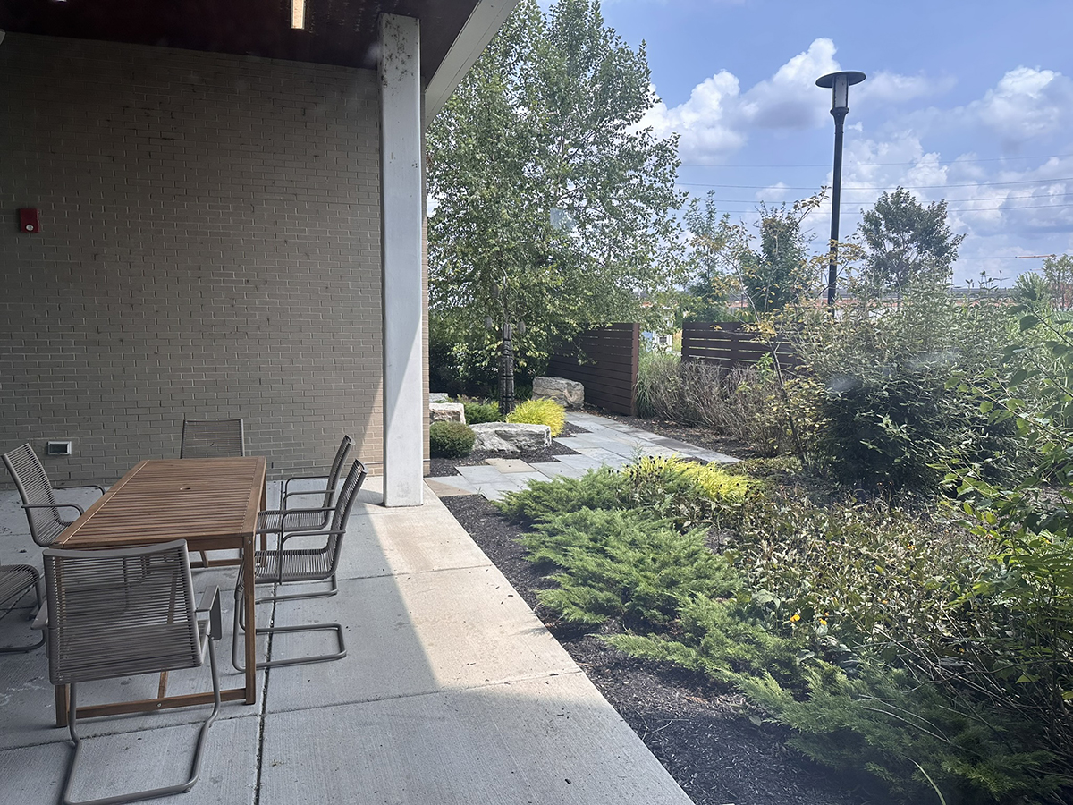 Outdoor patio with a wooden dining table and metal chairs beside a brick wall, overlooking a landscaped garden and stone path under a sunny blue sky.