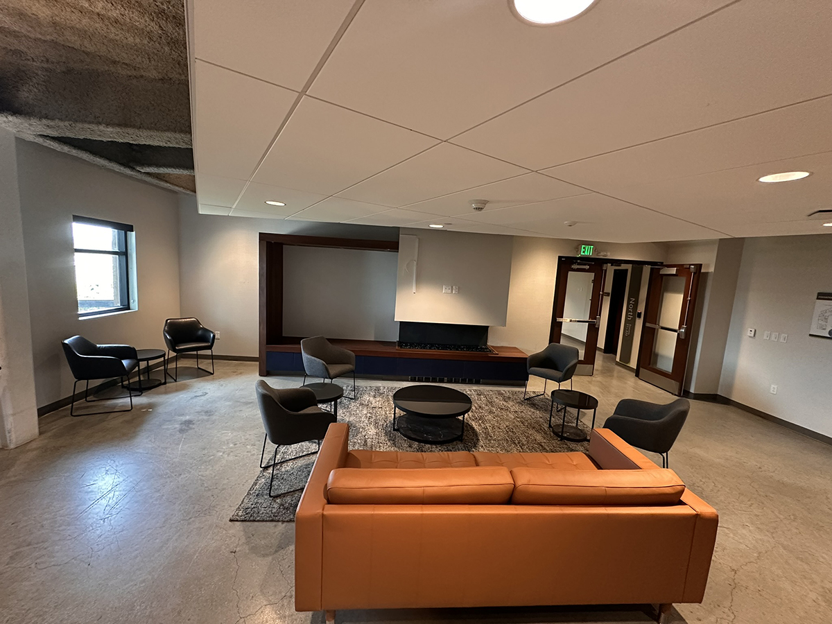 Modern office lounge with orange sofa, gray chairs, and round coffee tables around a rug, near a large screen wall unit.