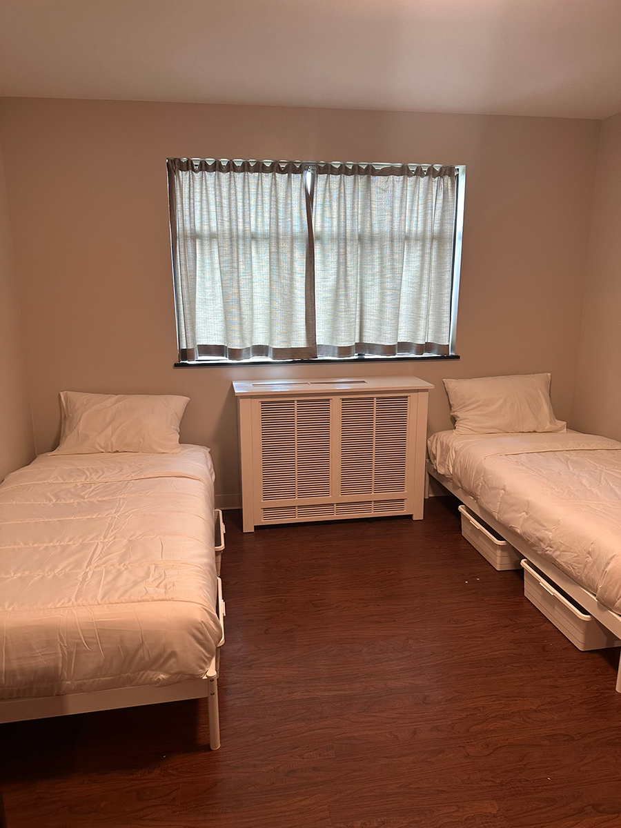 Twin bedroom with two single beds, white bedding, and a central radiator cover beneath a window with light curtains.
