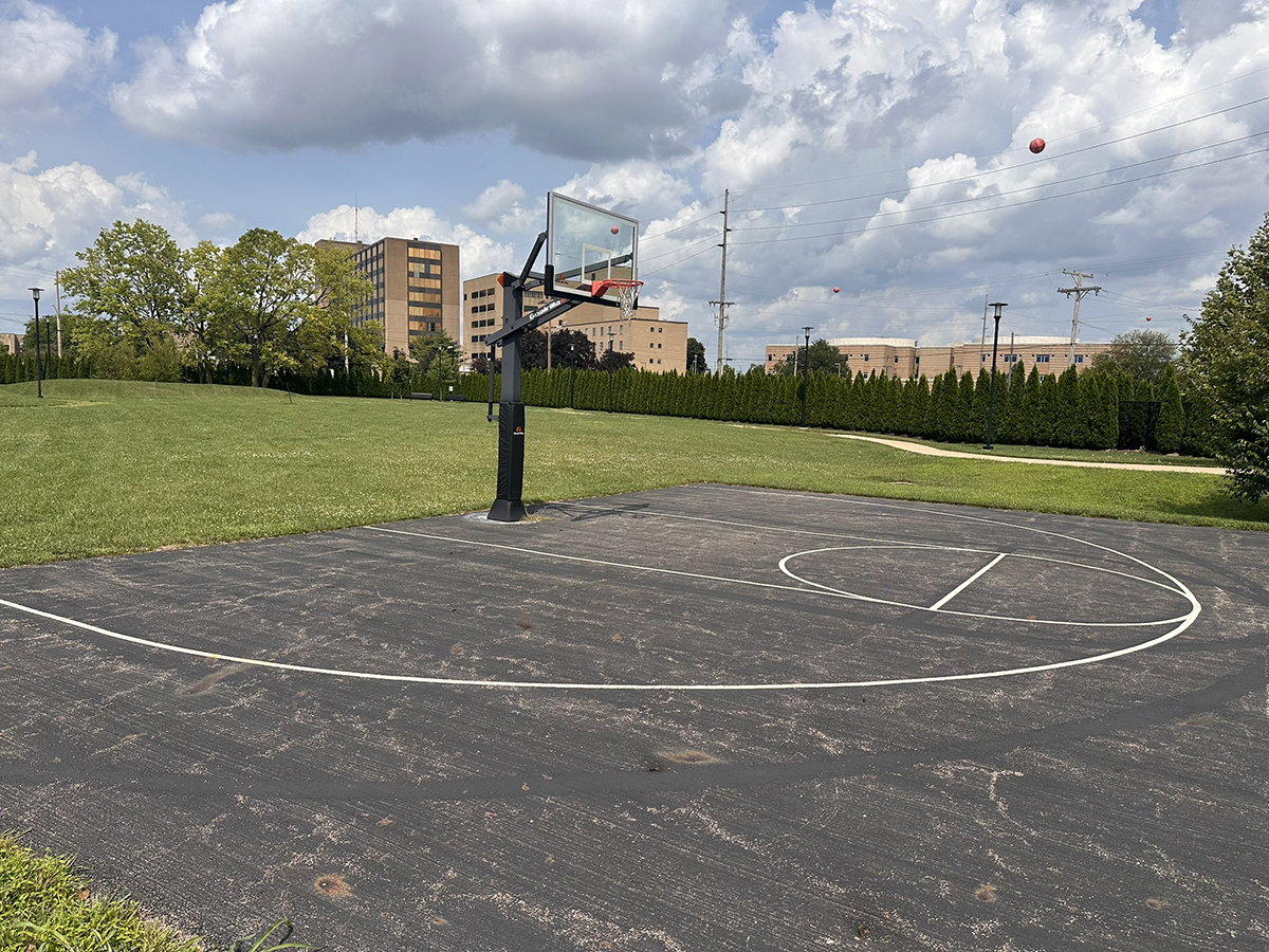 Outdoor basketball court with a backboard and hoop, surrounded by grass and trees, with buildings in the distance under a cloudy sky.